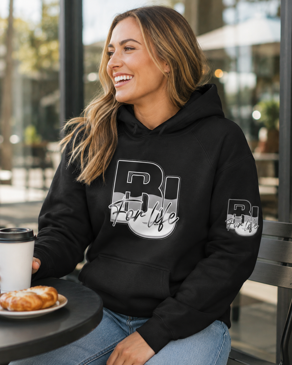 A woman smiling while wearing a black BU hoodie with a large white and gray 'BU For life' graphic on the front and a smaller matching graphic on the sleeve, seated at a table with a coffee cup and pastry in a cafe setting.
