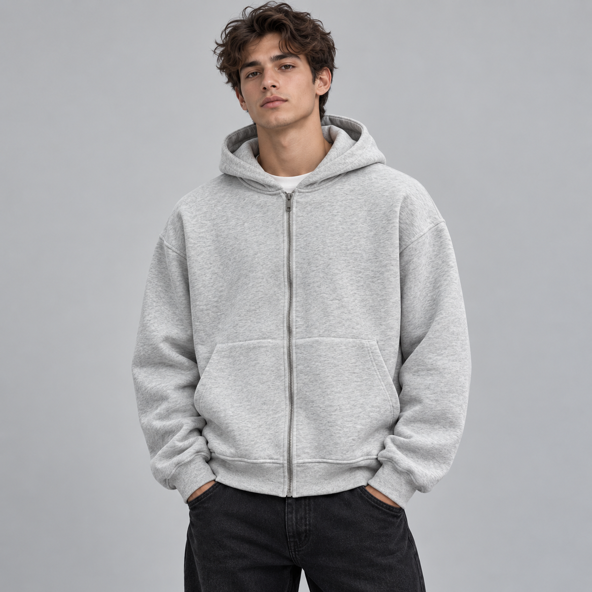 A young man wearing a light gray Hoodie Backpack  zip-up hoodie with a front kangaroo pocket, paired with dark jeans, standing against a solid gray studio background