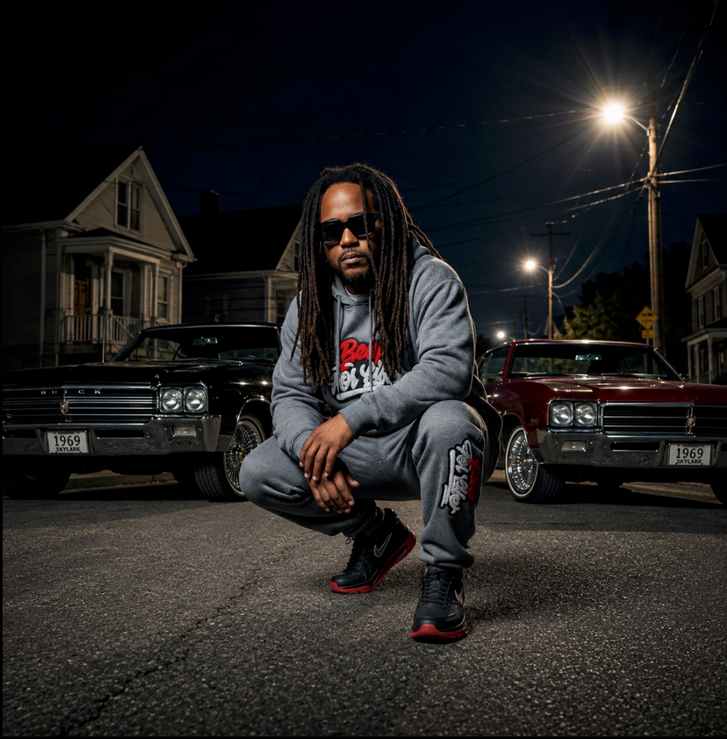 QGODFEAR wearing a gray BE UP FOR LIFE chenille patch jogger set crouching between two classic 1969 Buick Skylarks at night.